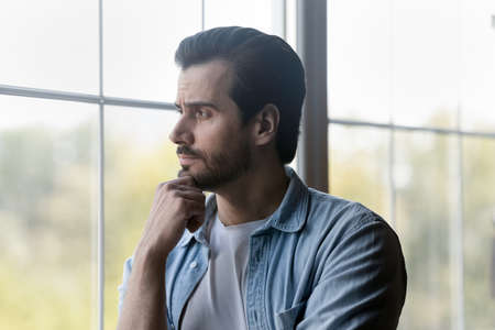Lost in thoughts. Pensive millennial bearded guy stand by window look at distance rub chin...