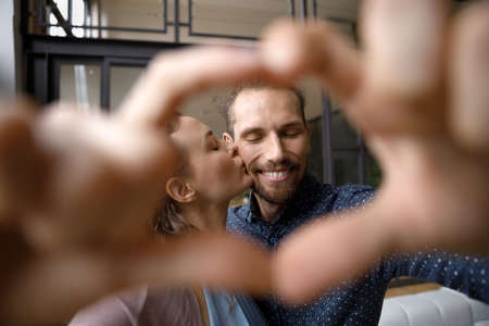 Head shot close up happy young woman kissing affectionate husband, making heart symbol with fingers. Loving millennial candid family couple showing sincere feelings at home, relations concept.の写真素材