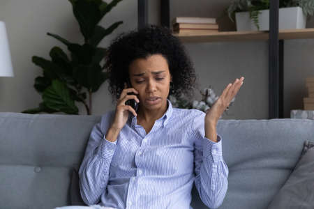 Unhappy African American woman talking on phone at home, sitting on couch, dissatisfied upset young female having unpleasant conversation, discussing problems, hearing bad news, complainingの写真素材