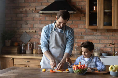 Helping daddy at kitchen. Tween child son learn to cook easy vegetarian salad watching young dad cutting ingredients on kitchen table. Preteen boy imitate father in preparing homemade food. Copy spaceの写真素材