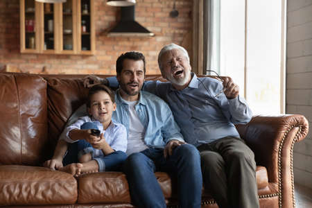 Energetic fans. Old grandpa young dad tween boy son sit on cozy sofa watch football game live broadcast on television. Three male members of multi generational family support soccer team on tv screenの写真素材