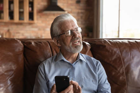 Thoughtful old age man sit on couch hold phone look aside distracted from screen recall in memory...
