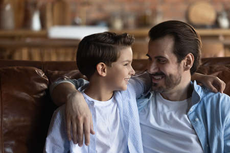 Pleasant talk with daddy. Affectionate single dad enjoy chatting with little son tell about passed day. Caring young foster father sit on couch embrace adopted child boy listen news from school lifeの写真素材