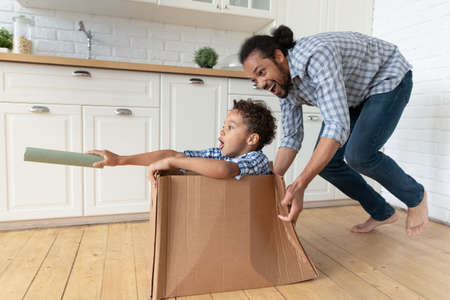 Happy Black dad driving cardboard box with excited little son inside, playing with kid at home, enjoying active games, leisure, entertainment after moving into new apartment. Family, relocationの写真素材