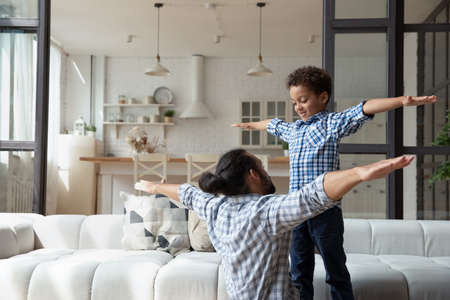 Happy cute African boy and dad enjoying active games, exercises at home, teaching yoga, playing airplane with open flying hands. Father and little son practicing fit activities, having fun togetherの写真素材