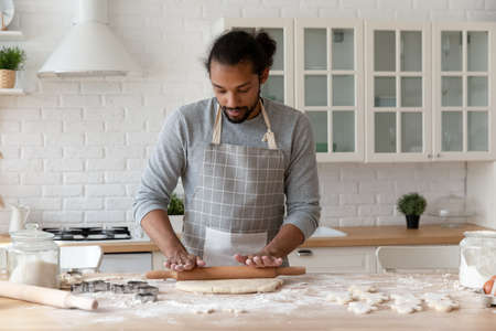 Focused young African American man baking cookies in home kitchen. Food blogger, baker, chef in apron rolling dough for biscuit, bread, cake, cooking bakery dessert at flour table with ingredients.の写真素材