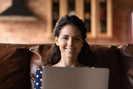 Having fun online. Smiling latin female laptop user rest on couch at home make video call watch funny photo content at social media. Positive young woman read good news online flirt at dating web chatの写真素材