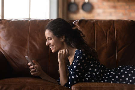 Yes. Happy cheerful millennial latin woman receive love message from boyfriend at phone messenger app. Excited young lady lying on sofa at home with cell read positive news celebrate achieving successの写真素材