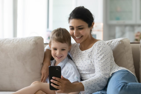 Happy mom and son making video call on smartphone, taking selfie, watching online content on mobile phone, relaxing and hugging on couch, looking at cell screen, smiling, laughingの写真素材