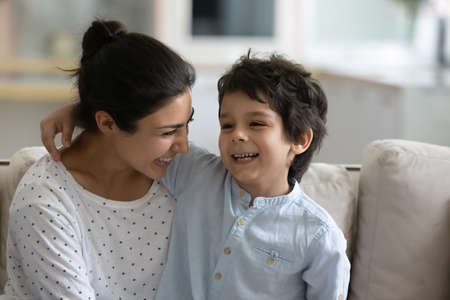 Happy cute Indian little son hugging laughing mom, sitting on lap, speaking, telling funny story. Millennial mother holding boy in arms, enjoying leisure with kid, cuddling child. Motherhood conceptの写真素材