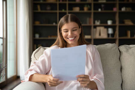 Happy young beautiful woman feeling pleased reading paper correspondence letter with good news, getting last bank mortgage payment notification or taxes refund, sitting on cozy couch at home.の写真素材