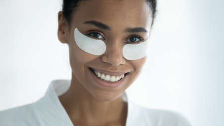 Close up head shot happy millennial african ethnicity multiracial woman with hydrogel moisturizing patches on under eyes area looking at camera, domestic self care, professional effective cosmetics.の写真素材
