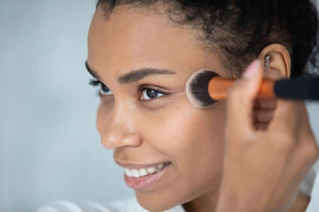 Close up smiling beautiful young african american mixed race woman applying foundation bronzer, contouring face using brush, getting ready doing daily nude make up, luxury products advertisement.の写真素材