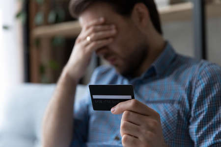 Depressed unhappy young man holding credit card in hands, feeling stressed having financial...