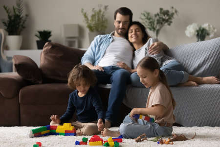 Two little kids together while parent relaxing on comfortable couch at home, smiling mother and father hugging, sitting on sofa, adorable sister and brother having fun, family enjoying leisure timeの写真素材