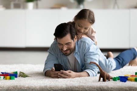 Smiling little daughter lying on father back, using smartphone together, having fun on warm floor at...