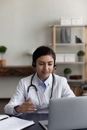 Vertical photo of young Indian woman doctor in headphones talking, using laptop in office, female physician general practitioner consulting patient online by video call, intern watching webinarの写真素材