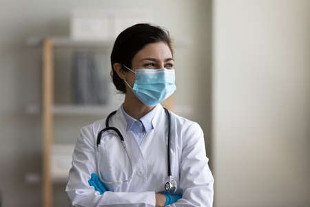 Happy Indian woman doctor in protective medical mask dreaming, visualizing and planning, looking in distance, smiling young female physician nurse in uniform with stethoscope standing in officeの写真素材