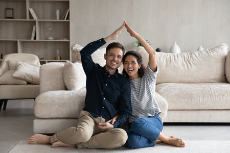 Happy Hispanic 35s couple joining hands under heads showing roof symbol sit on warm floor in fashionable living room. Homeowners spouses at relocation day, bank loan for young family, tenant conceptの写真素材
