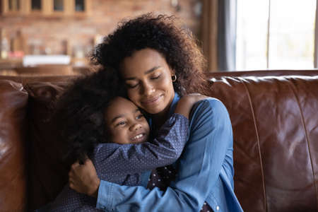 Happy loving African American little child girl cuddling young biracial mother, missing after long separation time, showing sweet tender feelings resting together on sofa at home, candid relations.の写真素材