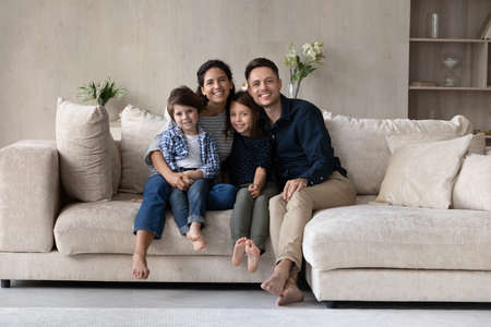 Beautiful young 35s couple and little children sit on cozy couch in living room smiling looking at...