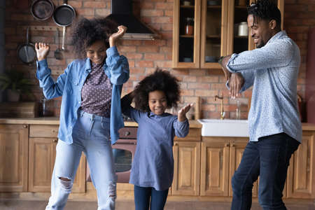 Happy laughing African American young parents jumping dancing to disco music, entertaining having fun with adorable small biracial kid daughter in kitchen, celebrating moving into own dwelling.の写真素材