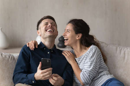 Hispanic couple sit on sofa in living room with smart phone, laughing over online videos, watch prank on internet looking overjoyed spend time at home use virtual amusement and entertainment conceptの写真素材