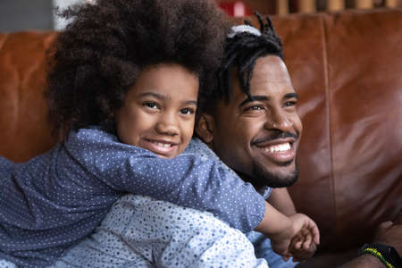 Happy little cute African American child girl embracing young Brazilian father, resting together on cozy sofa, enjoying stress free weekend time at home, good family relations concept.の写真素材