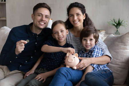 30s couple and little kids sit on sofa with piggy bank smile look at camera, save money for future, children education, investment for own house manage family budget, make savings for tomorrow conceptの写真素材