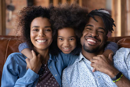 Head shot portrait of loving little kid girl cuddling young parents, showing tender feelings. Happy diverse African American family enjoying sweet moment at home, good relations concept.の写真素材
