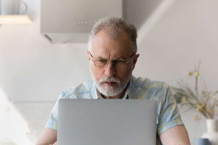 Focused older 60s man wearing glasses, working at laptop in home kitchen, typing, looking at screen....