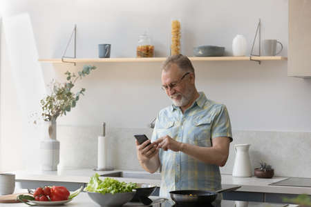 Cheerful older homeowner man consulting internet on smartphone, reading recipe, food blog online, cooking lunch in home kitchen, using gadget at table with fresh salad vegetables ingredientの写真素材