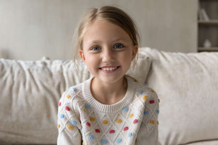 Head shot portrait of smiling adorable little kid girl sitting on comfortable couch, holding...