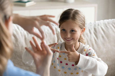 Smiling cute little preschool child girl with hearing disability making gesture with fingers, learning sign language with professional female therapist or practicing communication with young mother.の写真素材