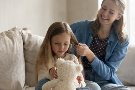 Happy cute small kid girl playing with favorite toy while caring affectionate mother combing her blonde light hair, getting ready for kindergarten or school, doing hairstyle, sitting on couch at home.の写真素材