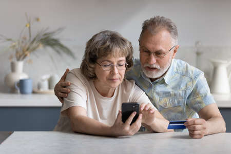 Serious older married couple shopping on internet stores, using banking, ecommerce app on cellphone, paying for purchase by credit card, making transaction, transfer, money order. Finance conceptの写真素材