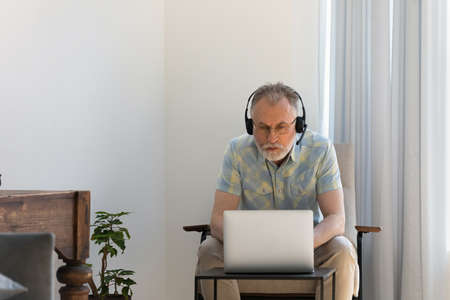 Focused older 60s man in wireless headset using laptop at home. Male online teacher, coach giving webinar, training, lecture, learning course, making video call, sitting in armchair at computerの写真素材