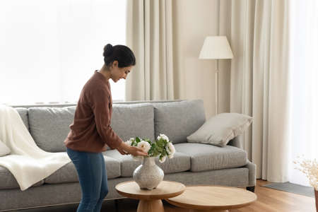Smiling young indian ethnicity woman homeowner putting beautiful flowers in vase on coffee table in modern living room, enjoying decorating own apartment, improving styling interior on weekend.の写真素材