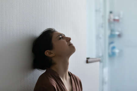 Head shot side view stressed frustrated young indian ethnicity woman resting head against wall, feeling depressed alone at home, suffering from negative thoughts or mental disease, crying indoors.の写真素材