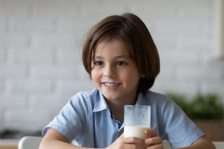 Lovely 6s boy with milk moustaches holding glass of dairy natural beverage sit alone indoor smiling looks into distance. Balanced diet for children health and growth, healthy eating lifestyle conceptの写真素材