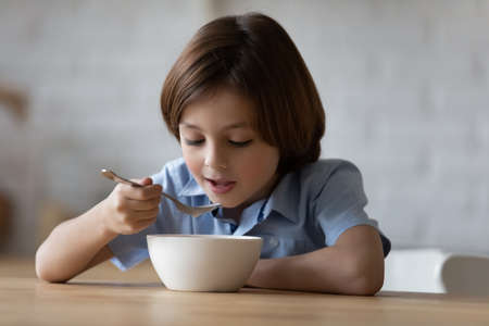 Little hungry cute boy sit at dinner table in kitchen holds spoon eating porridge or whole grain dry cereal with pleasure and appetite, enjoy balanced food. Nutrition, tasty healthy breakfast conceptの写真素材