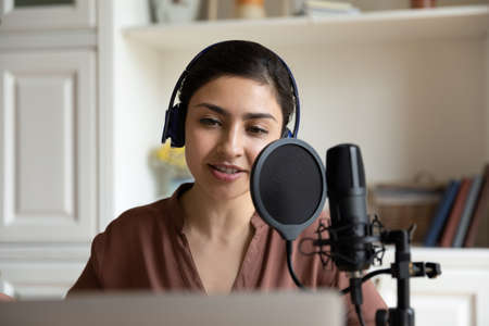 Head shot beautiful young indian ethnicity woman in headphones talking in professional stand microphone, recording audio for personal blog, singing song or voice acting using computer applications.の写真素材