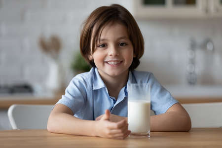 Cute little 6s boy sit at table in kitchen with glass of milk smiling looking at camera. Concept of balanced diet of children, delicious dairy calcium natural beverage, child growth, healthy lifestyleの写真素材