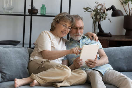 Happy elder 60s family couple with digital device relaxing on sofa together. Middle aged husband and wife using online app on tablet for internet shopping, virtual order, communicationの写真素材