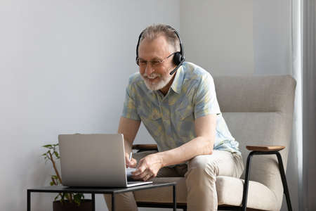 Happy mature online course student watching learning webinar, attending virtual class. Smiling senior man wearing headphones, using laptop at home, making video call, laughing, listening lectureの写真素材
