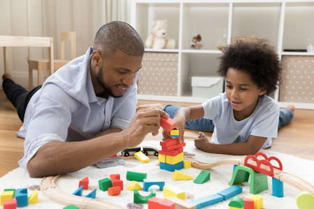 Loving young African dad and cute little son engaged in game with construction cubes, railway, resting and playing on warm floor, talking, enjoying playtime activities together. Family, fatherhoodの写真素材