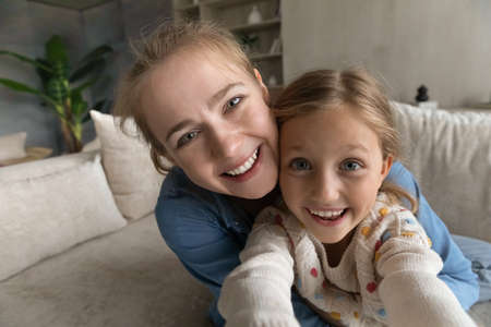 Close up sincere adorable little child girl having fun with happy loving mother, recording funny video or vlogging streaming stories online in social networks, posing for selfie photo, web camera viewの写真素材