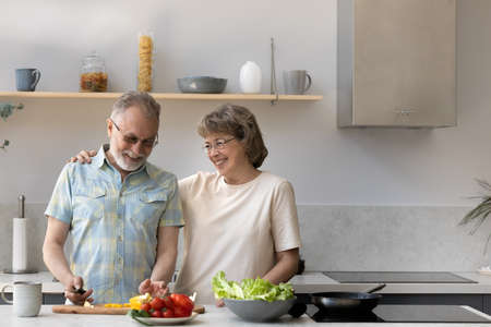 Happy joyful older husband and wife sharing cooking chores, making organic fresh salad together, cutting vegetables, talking, laughing. Old couple having fun in kitchen, preparing dinnerの写真素材