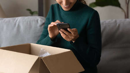 Positive feedback. Close up of young woman leaving comment at web store site after receiving parcel with goods purchased. Smiling millennial female order consumer products delivery online using phoneの写真素材