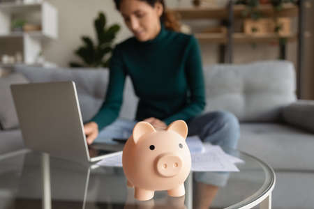 Wise economy. Blurred cropped shot of young woman managing household budget at home office planning family finances paying utility bills using app on laptop. Focus on funny pink piggybank toy on tableの写真素材